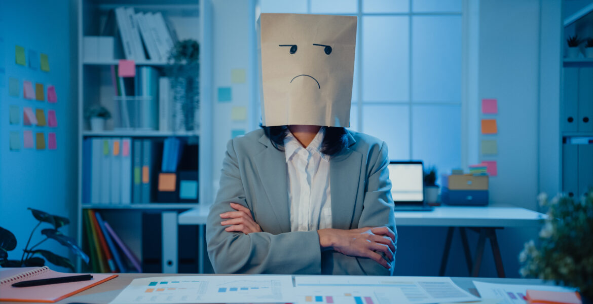 Young Asian businesswoman sit on desk wear draw sad mask paper craft bag feel bad mood with fail work project in office at night. Anonymous lady mad and confuse with overwork, Work mental health.