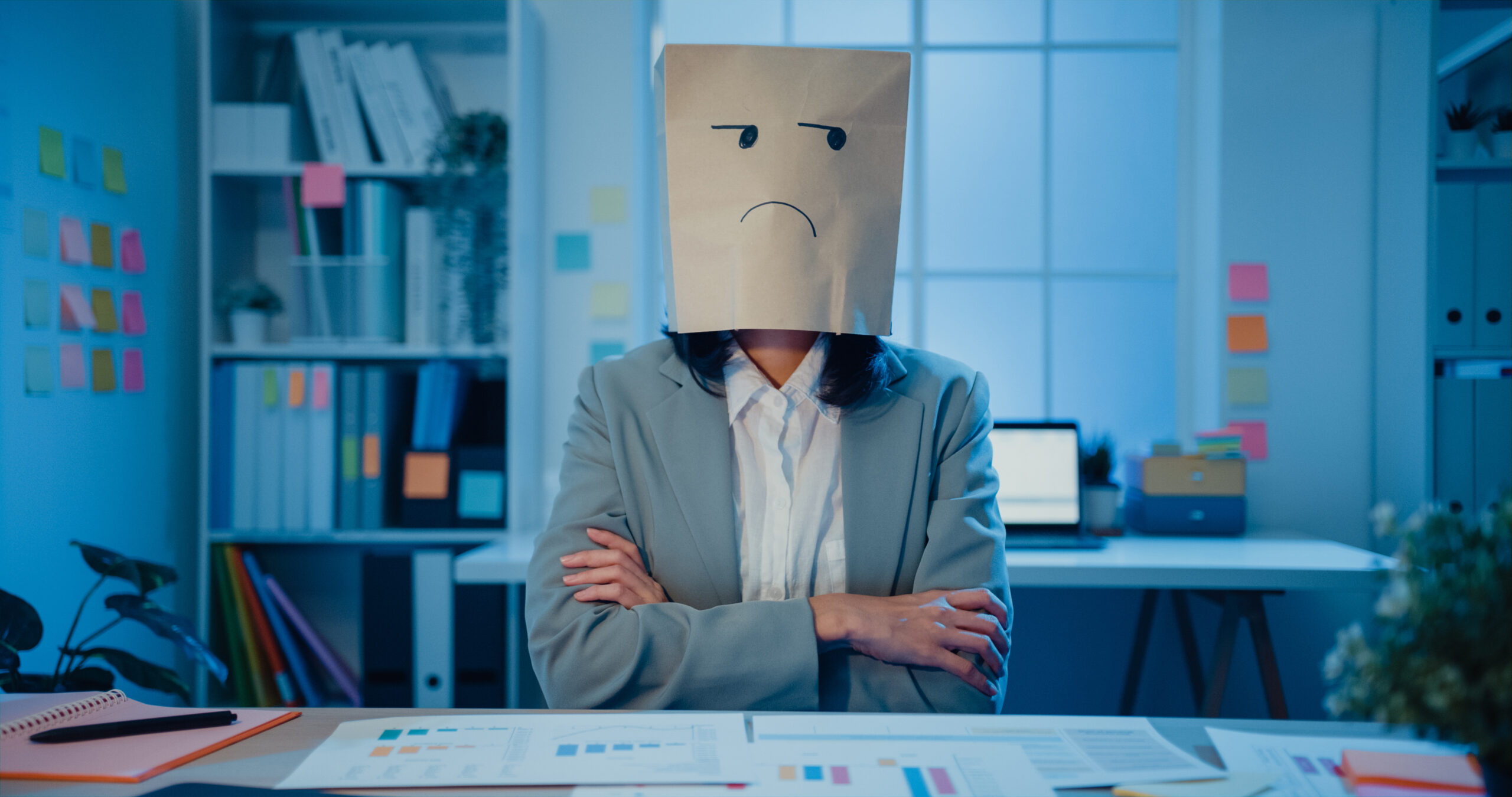 Young Asian businesswoman sit on desk wear draw sad mask paper craft bag feel bad mood with fail work project in office at night. Anonymous lady mad and confuse with overwork, Work mental health.